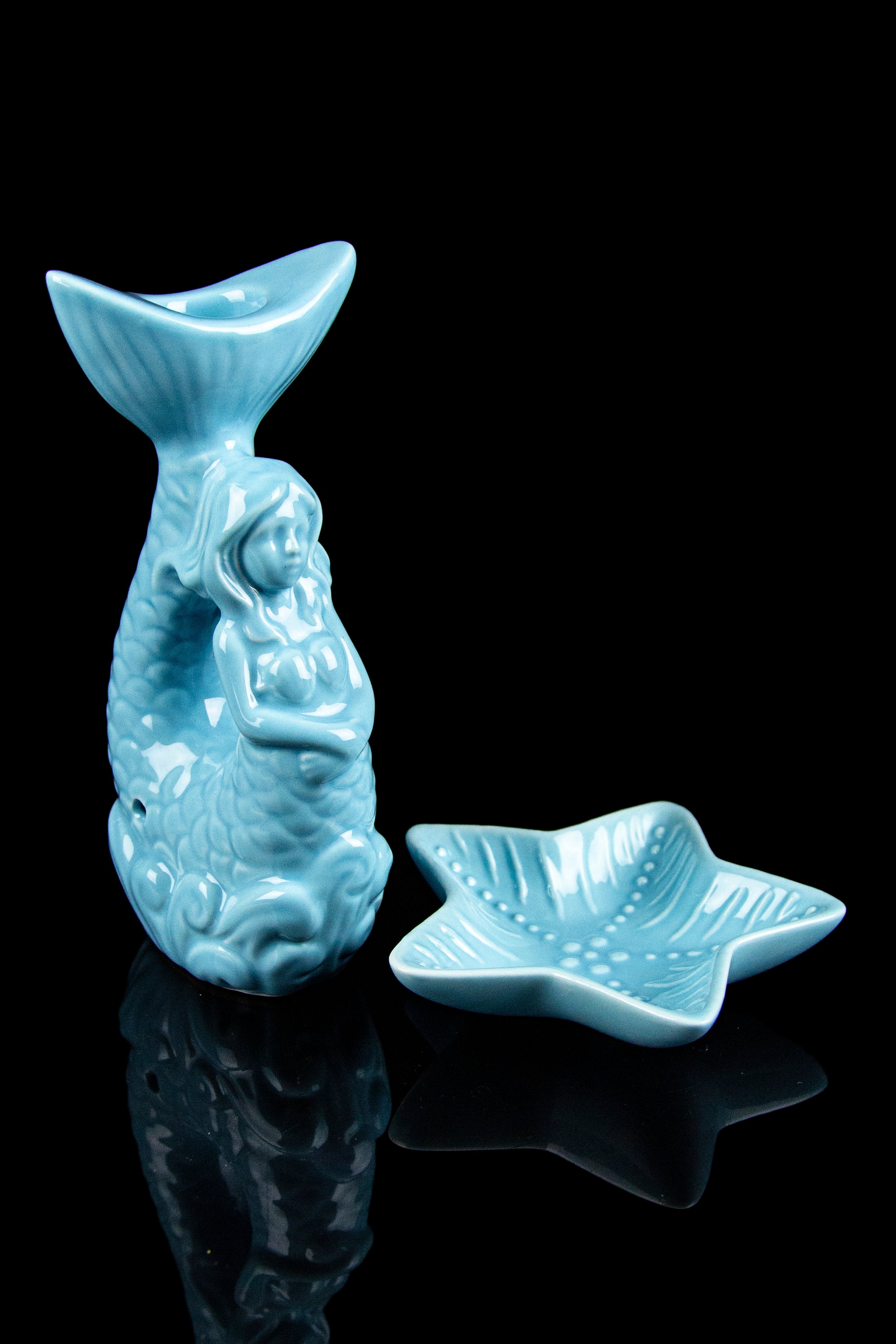 Ceramic mermaid-themed cannabis accessories, including a water pipe and ashtray, displayed on a reflective black surface.
