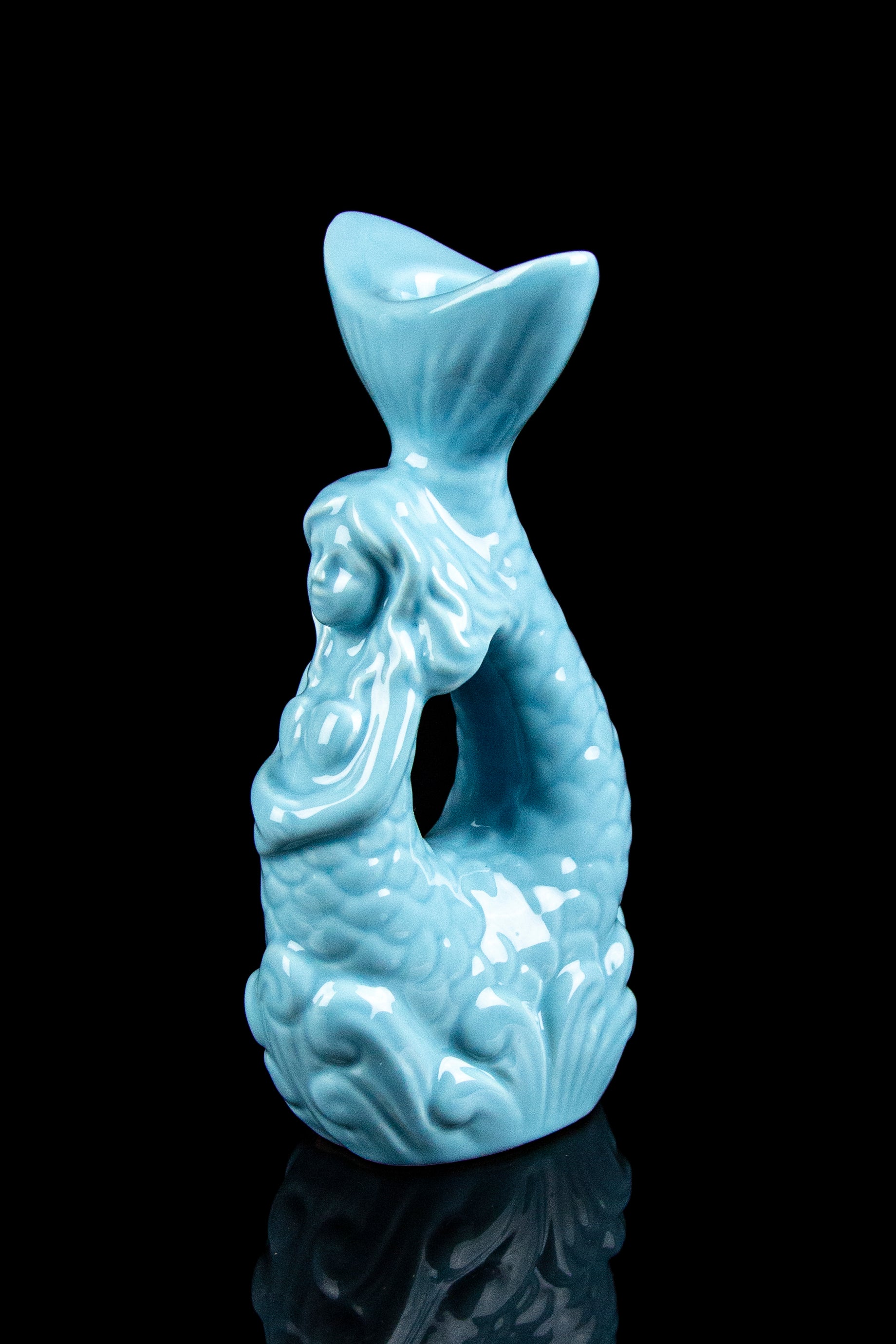 Turquoise glass sculpture of a mermaid, likely a water pipe or cannabis accessory, on a reflective black surface.