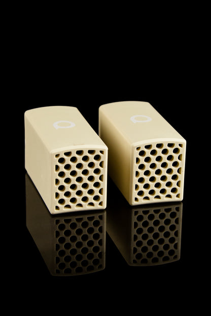 Two cream-colored ceramic cannabis filter tips with hexagonal perforations, reflecting on a black surface.