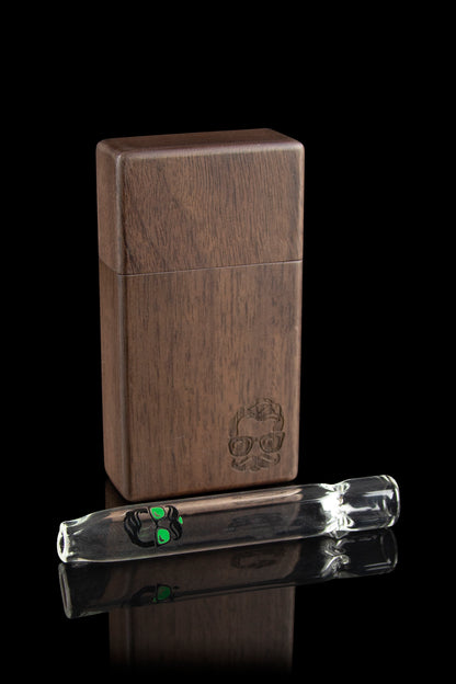 Wooden dugout with a glass one-hitter resting beside it, both featuring a green skull logo, against a black reflective surface.