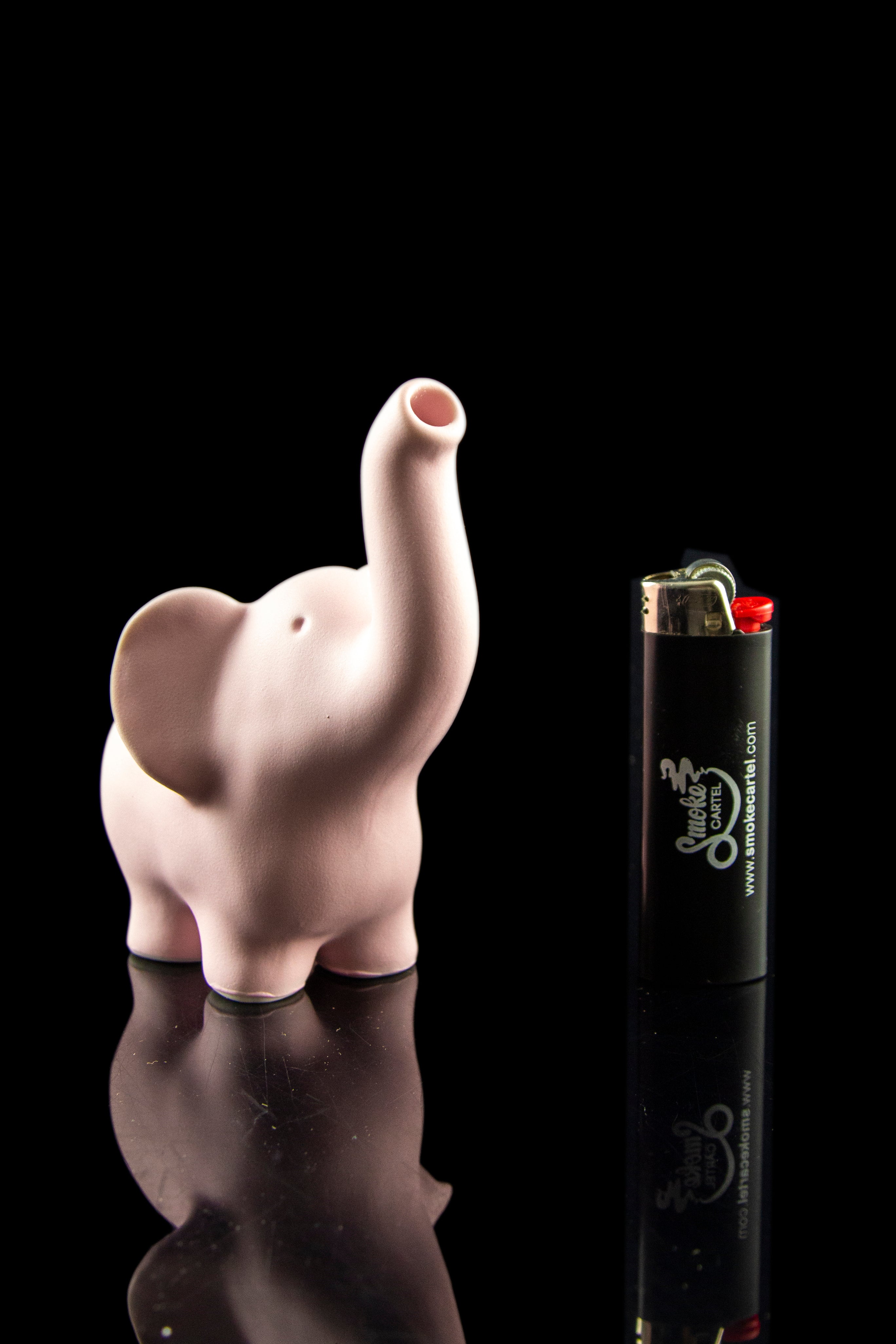 Pink glass elephant sculpture beside a Smoker's cartel lighter, reflected on a glossy black surface.