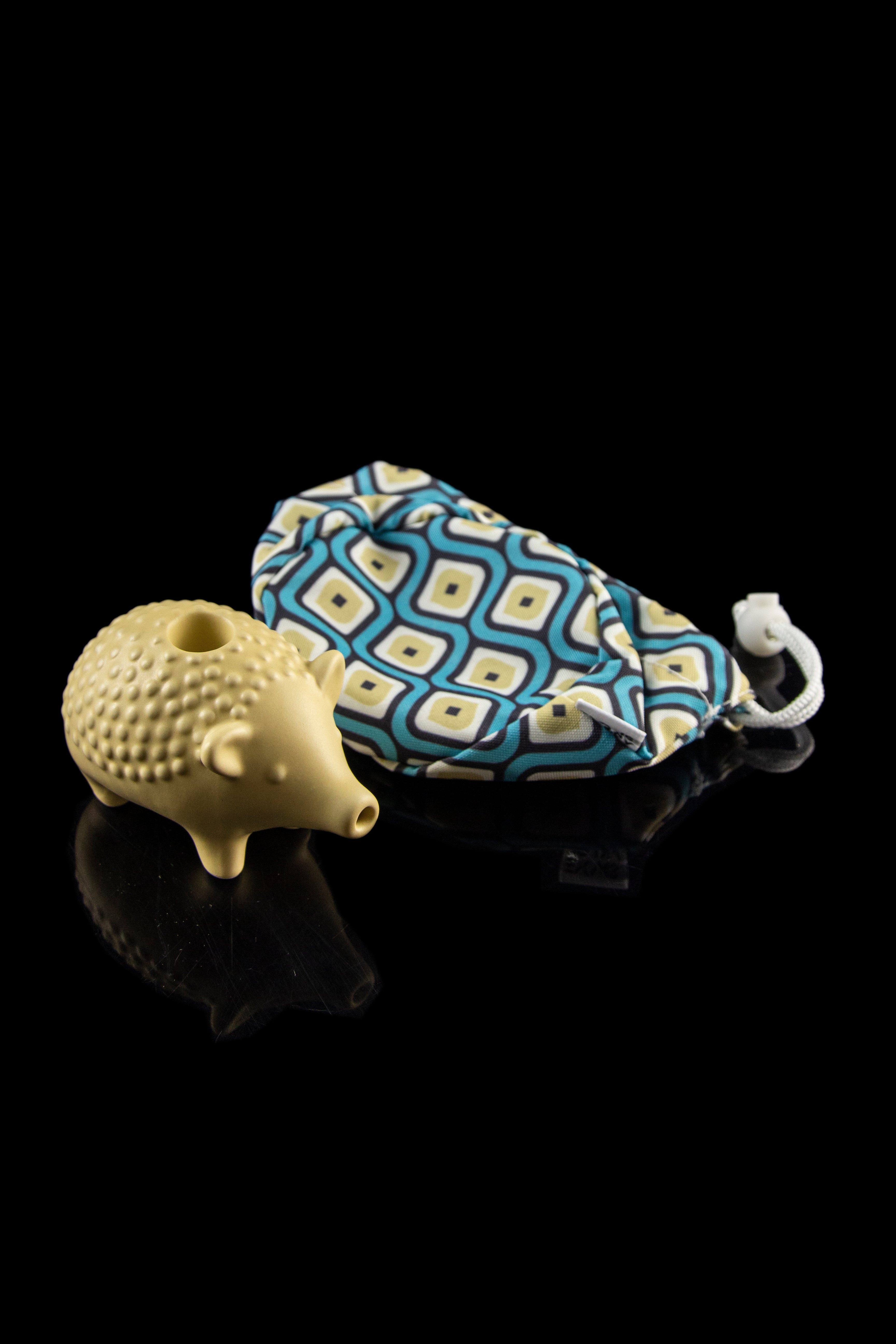A ceramic pipe shaped like a hedgehog rests beside a patterned cannabis travel case on a reflective surface.