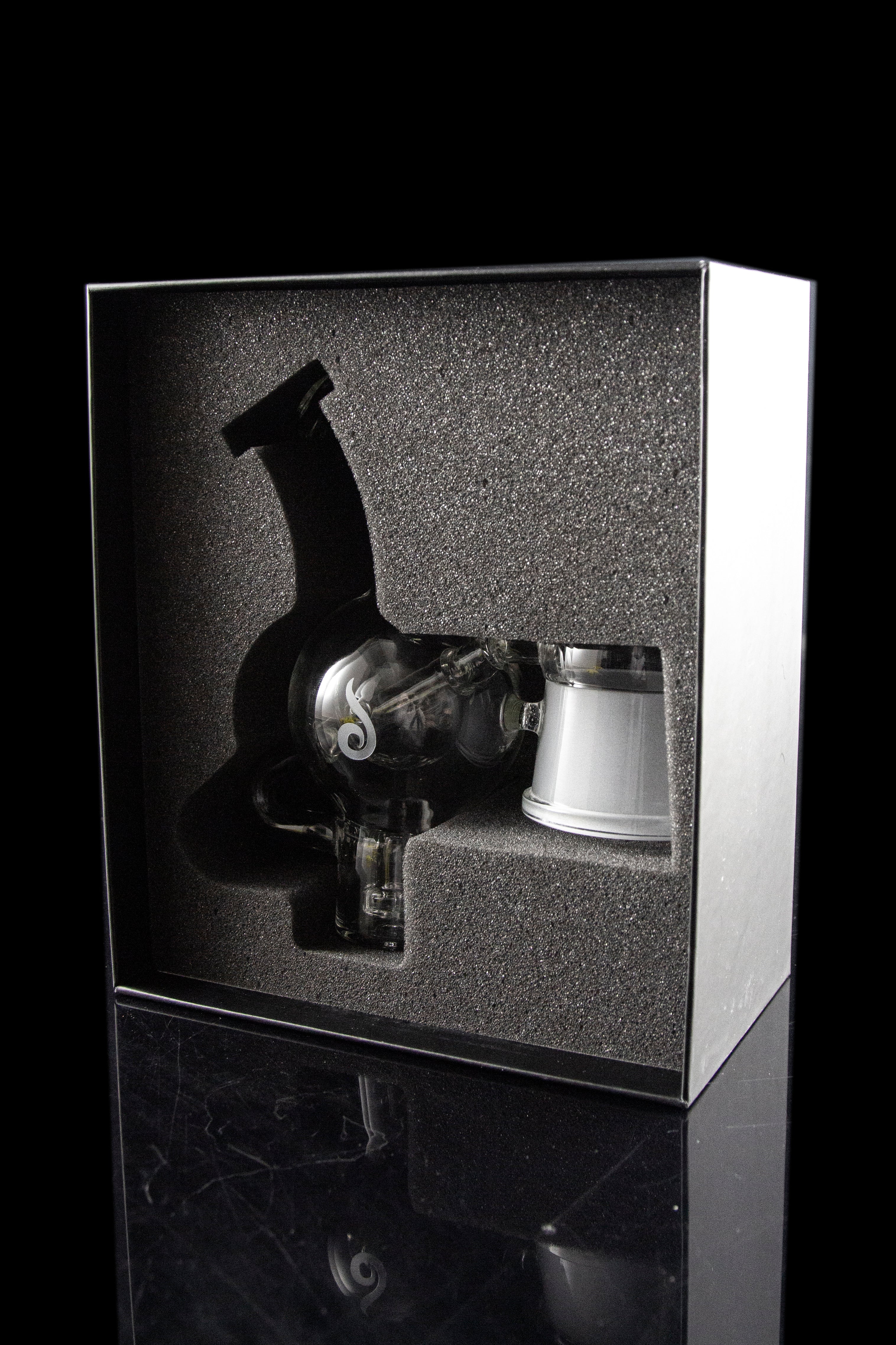 A cannabis dab rig and accessories displayed in a sleek, black presentation box with a reflective surface.