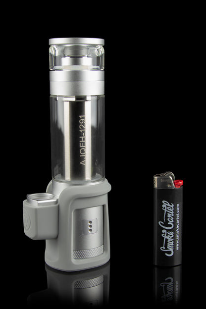 A silver and black electronic dab rig with a glass water pipe attachment, positioned next to a lighter with cannabis-related branding.