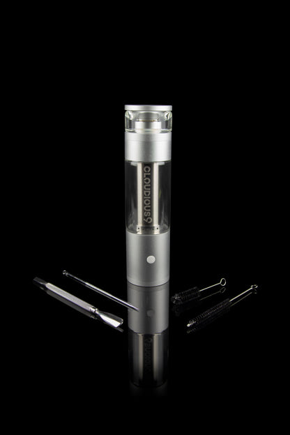 Silver cannabis vaporizer with glass chamber and cleaning tools on a reflective surface.
