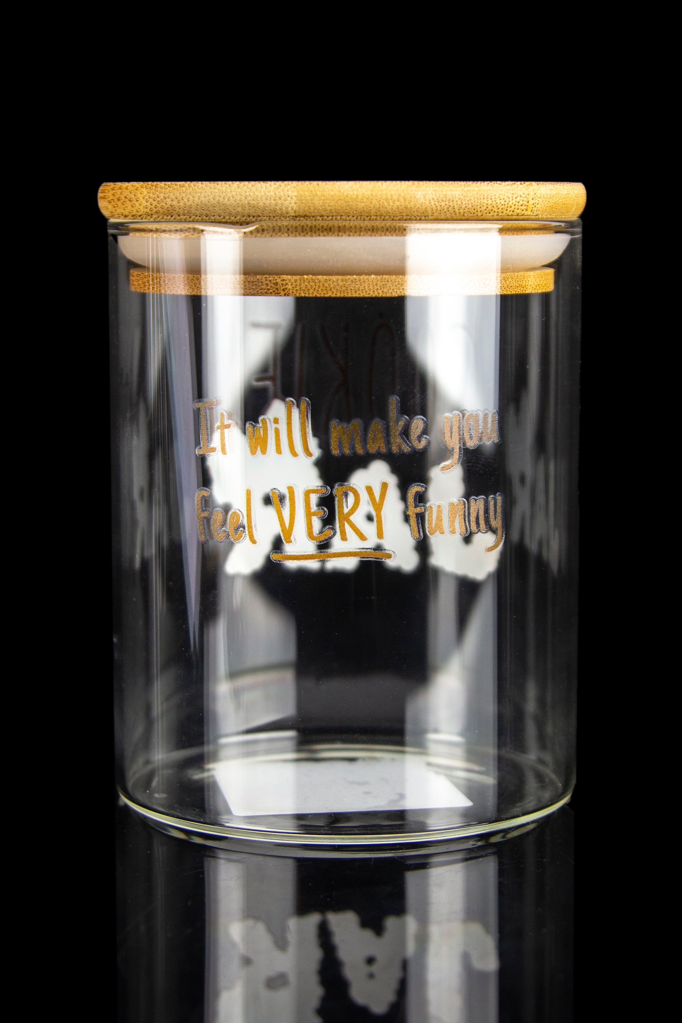 Clear glass jar with a bamboo lid and the text “It will make you feel VERY Funny” displayed on the front, reflected on a black surface.