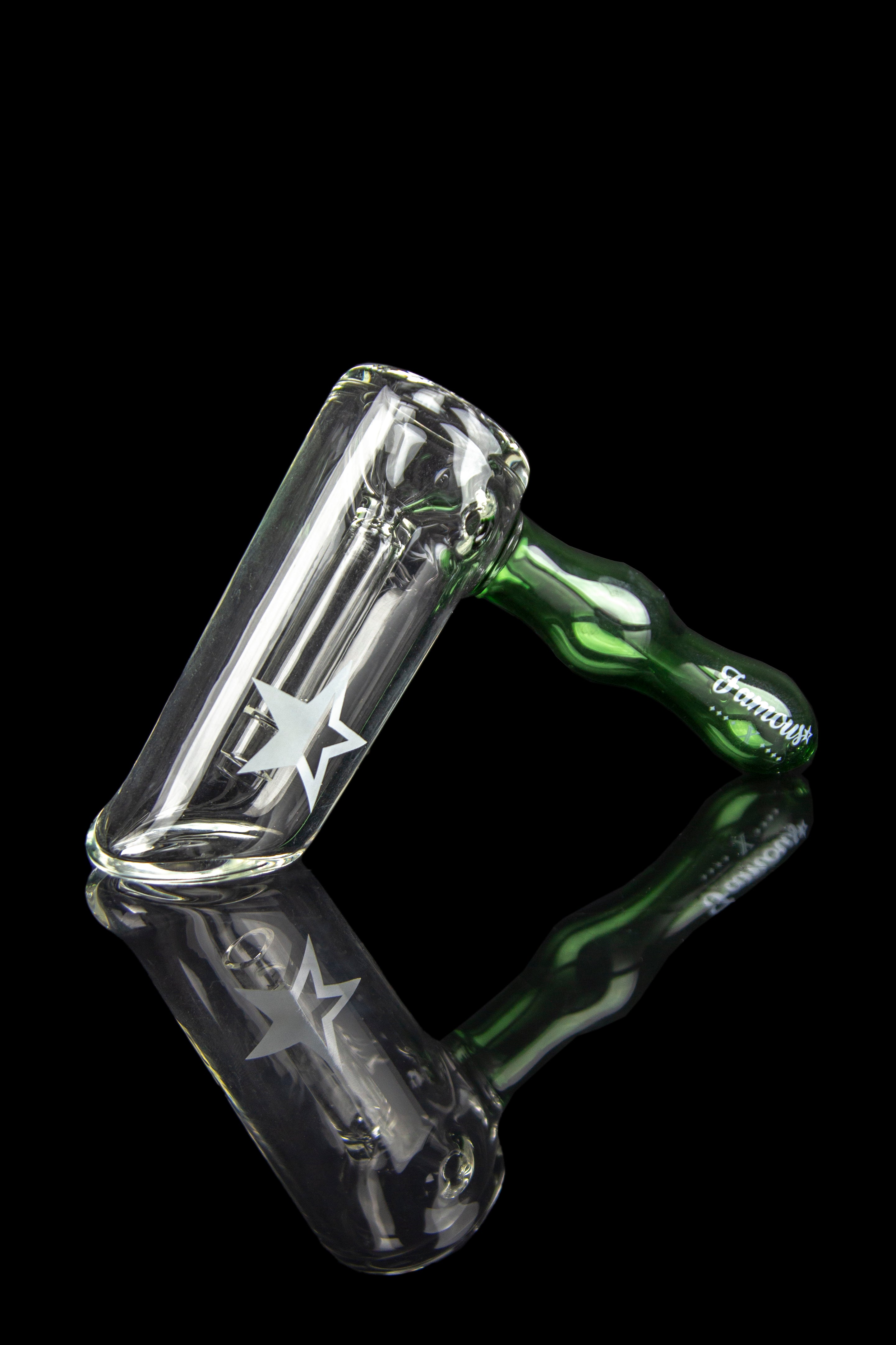Angled close-up of the Famous X Straight Hammer Bubbler Pipe showing its clear glass chamber with a white star decal and a green handle reflected on a black surface
