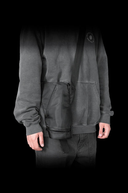 A person wearing a dark gray hooded sweatshirt and pants, with a cannabis accessories carrying case slung across their body.