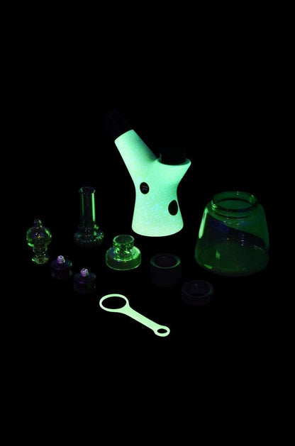 Glowing green glass cannabis water pipe and accessories on a black background.