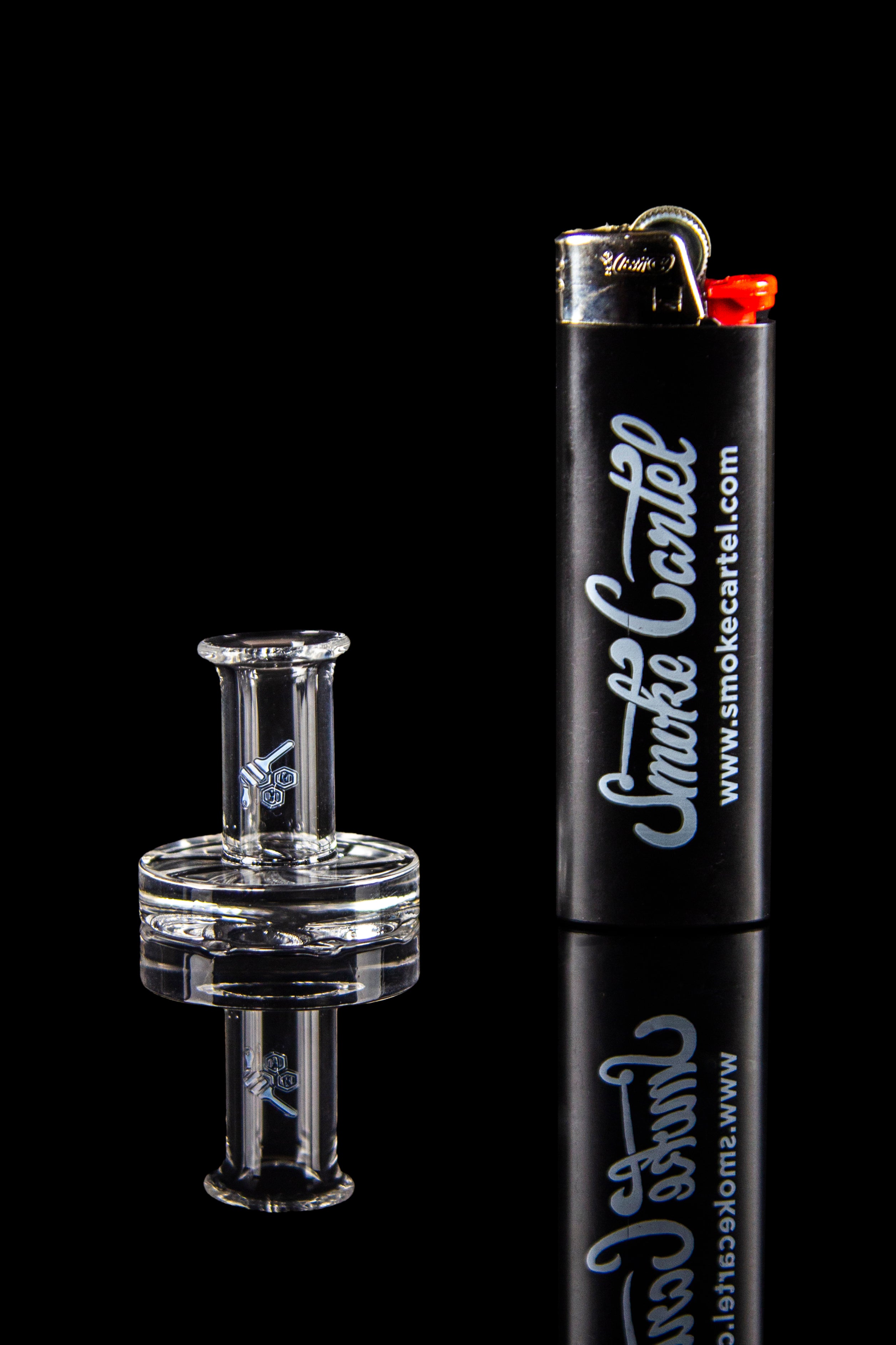 Clear glass cannabis oil rig with a black lighter branded “Smoke Cartel” reflected on a glossy black surface.
