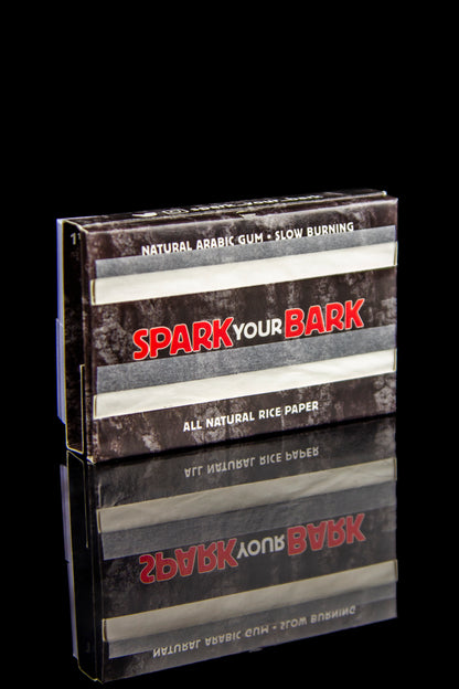 A pack of "Spark Your Bark" natural rice rolling papers with a woodgrain texture, set against a black background.
