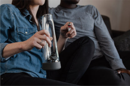 Two people seated on a couch, one holding a glass cannabis water pipe (bong) and the other holding a lighter."