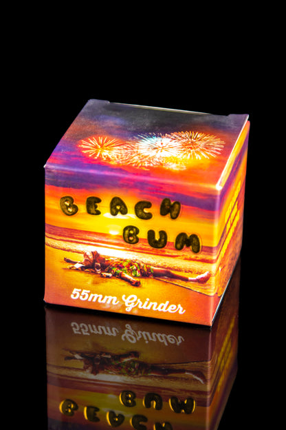 Box for a "Beach Bum" 55mm grinder with a sunset and palm tree design, reflecting on a dark surface.