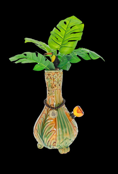 A vibrant, tropical-themed glass water pipe resembling a palm tree with a toucan perched among large green leaves, set against a black background.