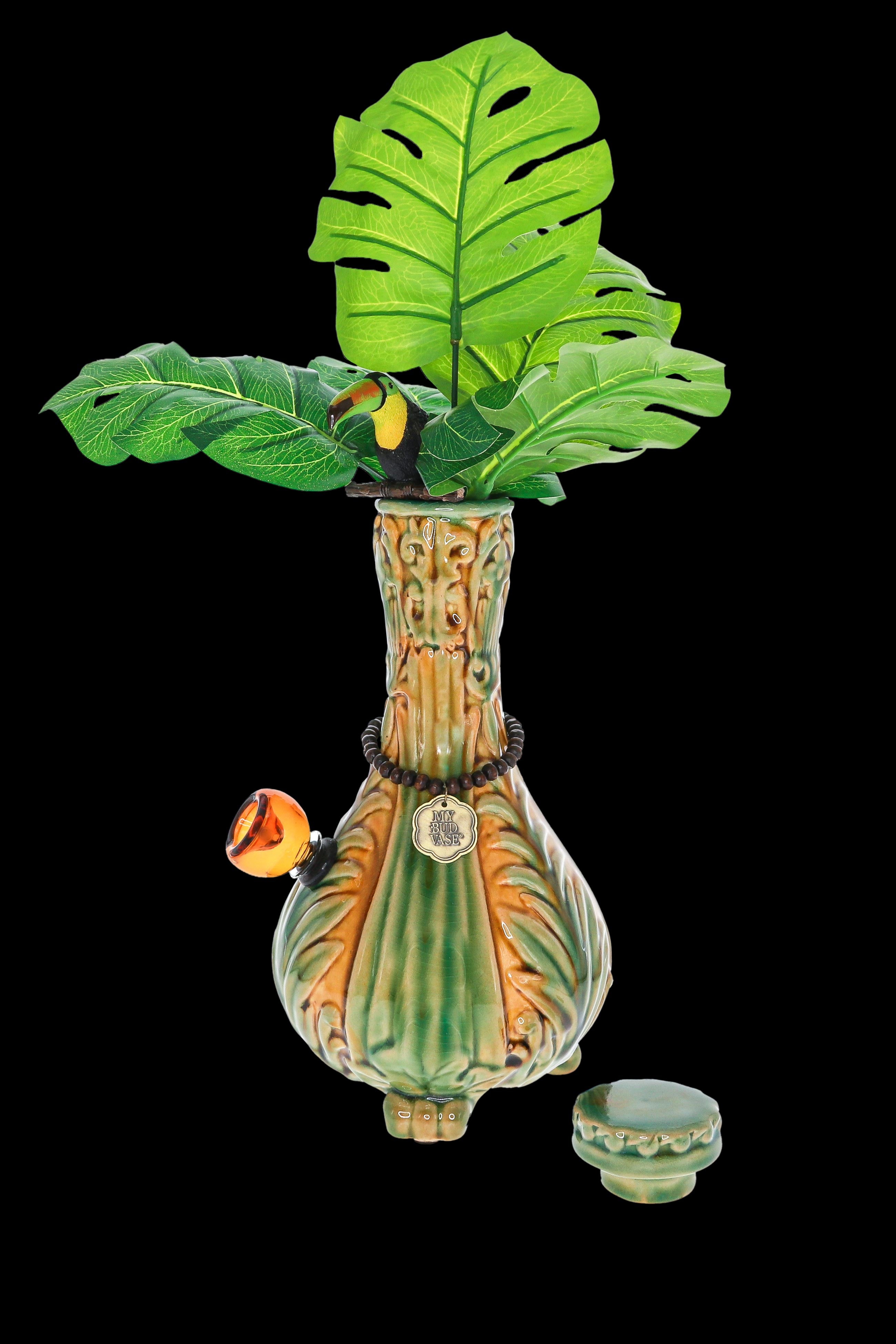 A vibrant, ceramic cannabis water pipe shaped like a tropical plant with detailed leaf and fruit accents, displayed with its accompanying bowl and base on a black background.