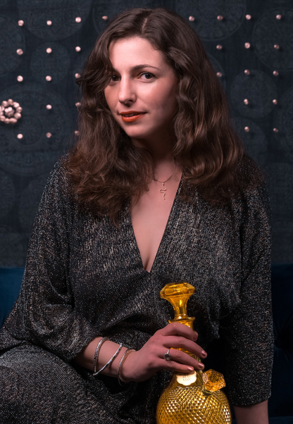 A woman with wavy auburn hair and a silver sequined dress holds a vintage, ornate yellow glass oil container, possibly for cannabis concentrates, against a dark floral backdrop.