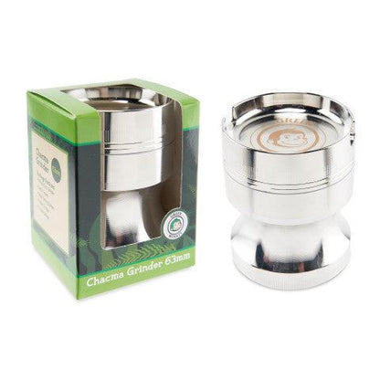 Two-item product shot showing the Green Monkey Chacma Grinder with Ashtray from a slightly elevated front angle, the silver metal grinder beside its green boxed packaging on a white background.