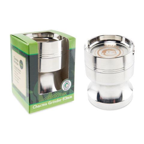 Two-item product shot showing the Green Monkey Chacma Grinder with Ashtray from a slightly elevated front angle, the silver metal grinder beside its green boxed packaging on a white background.