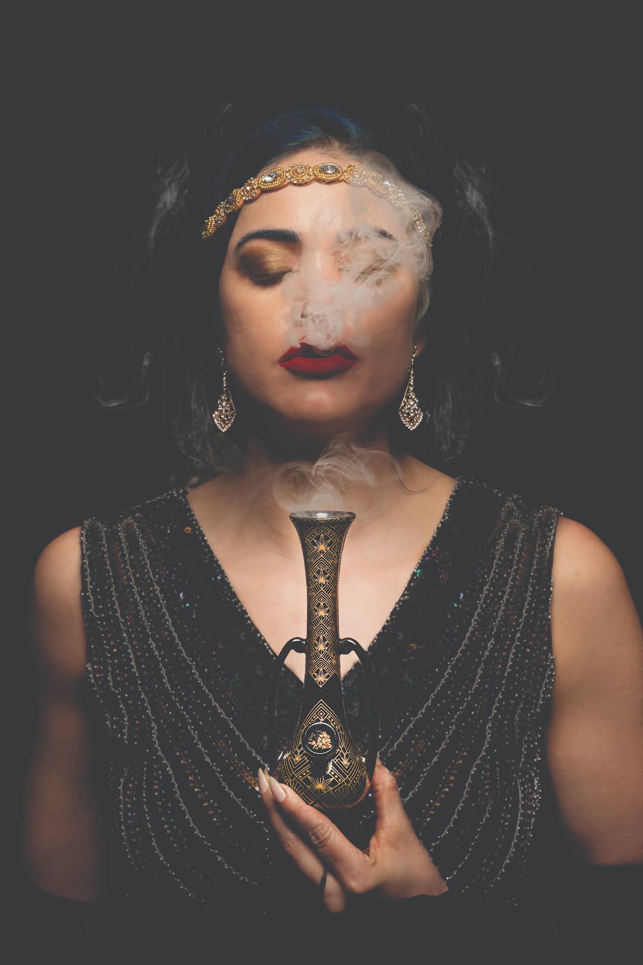 A woman with blue hair and dramatic makeup exhales smoke from a decorative glass pipe, wearing a beaded flapper-style dress and art deco jewelry against a black background.