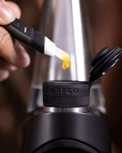 A hand applies cannabis concentrate to a Puffco Peak vaporizer using a dab tool.