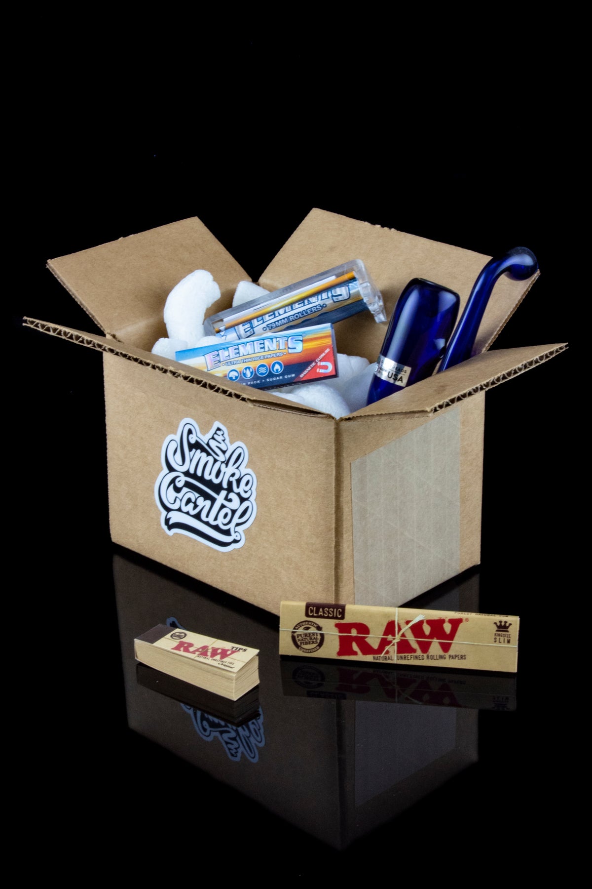 Open cardboard box filled with cannabis smoking accessories including papers, filters, and a blue glass pipe, displayed with branded packaging and reflected on a glossy surface.