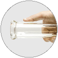 A hand holds the clear glass “Tank Glass “The Original” Tank Beaker” from a low angle, highlighting its cylindrical shape.